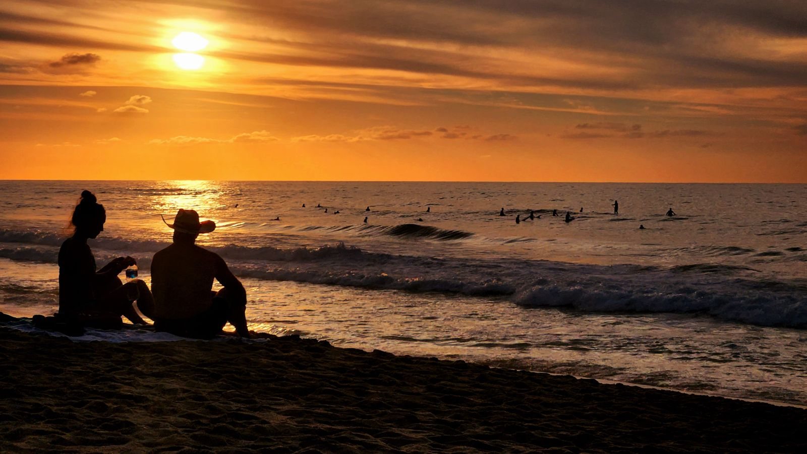 Silhouettes of a couple sitting on the sand at sunset while surfers wait for waves in San Pancho