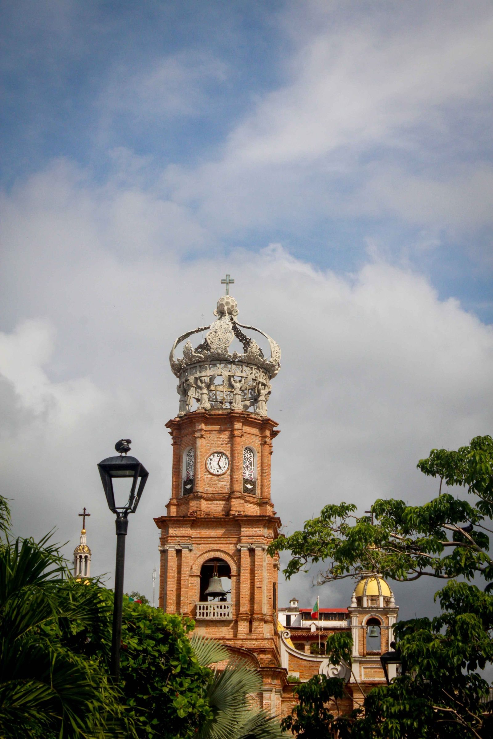 Tower of the Church of Our Lady of Guadalupe with its crown, bell and clock among trees and a street lamp