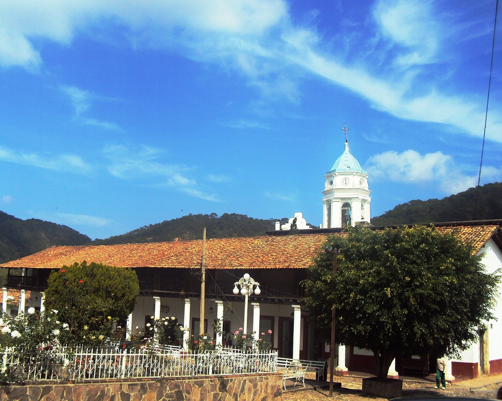 White bell tower of the San Sebastián parish above tile roofs, with forested mountains behind