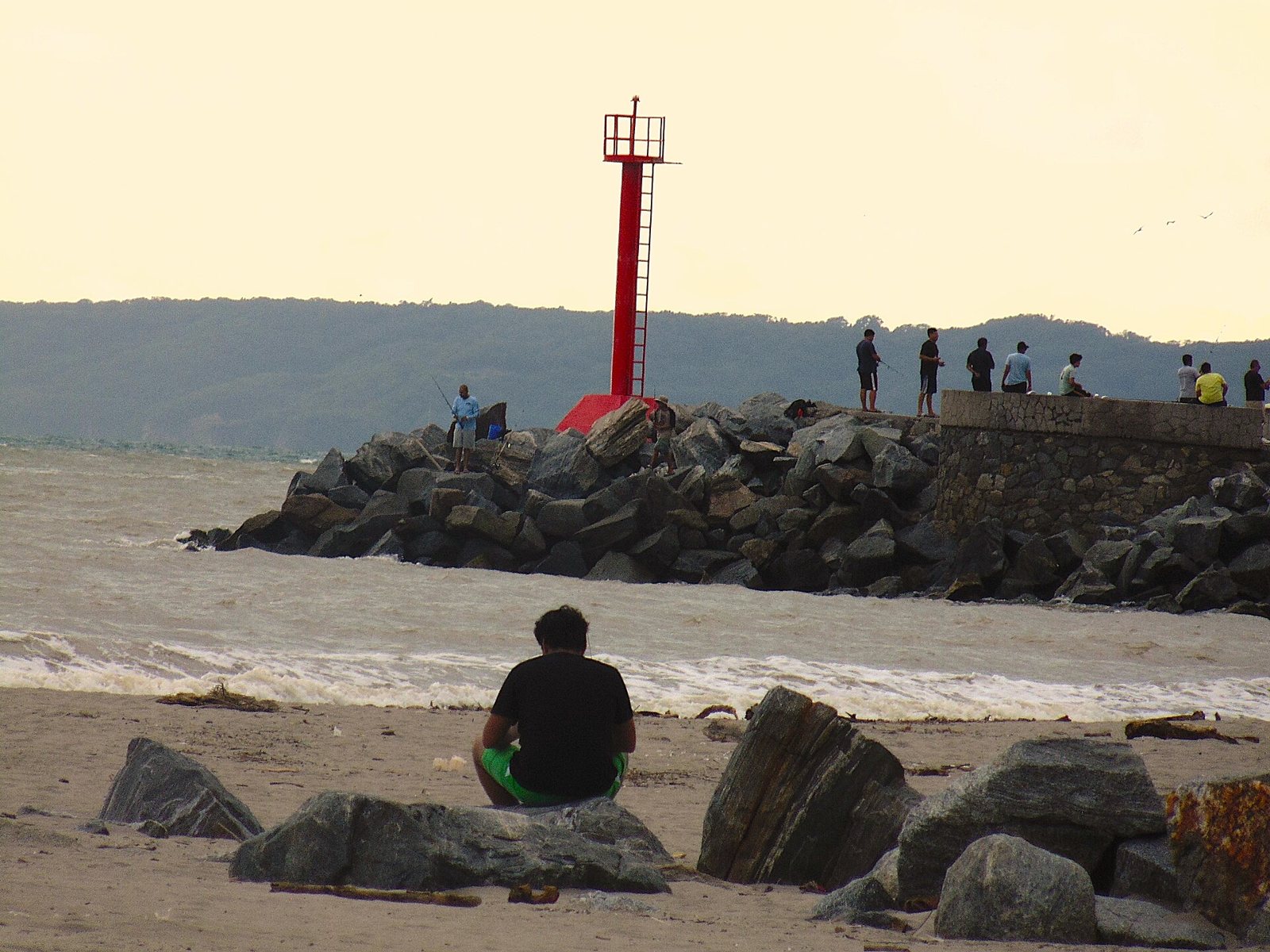 Red lighthouse on the marina jetty, with people fishing and mountains in the background