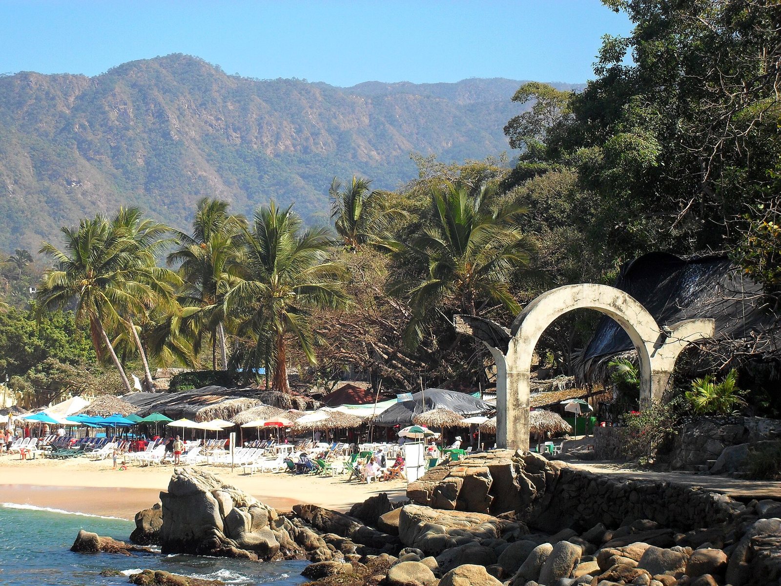 Palapas and loungers on the sand at Mismaloya, with palm trees, a stone arch and mountains behind