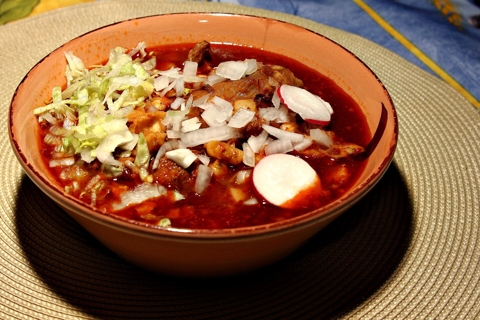 Bowl of red pozole with pork, hominy, shredded lettuce, chopped onion and radish slices