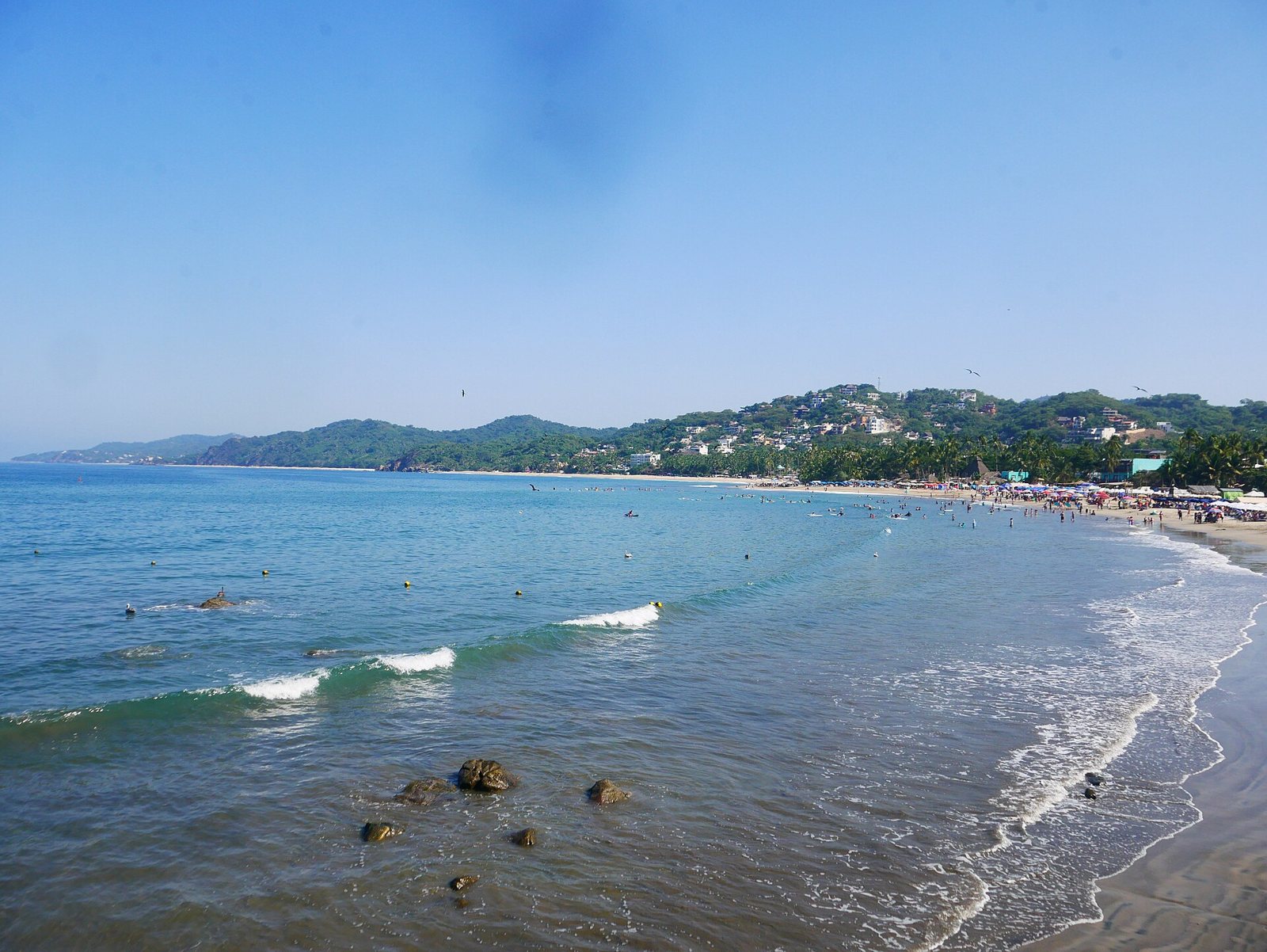 Small waves breaking across Sayulita bay, with swimmers and surfers in the water and the town behind