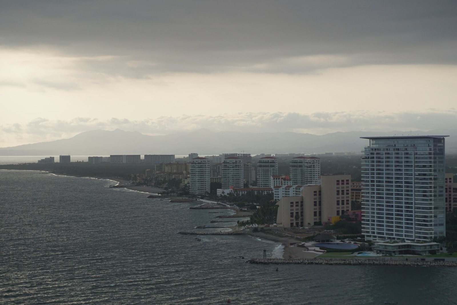 Banderas Bay shoreline with beachfront buildings and the silhouette of the Sierra Madre mountains under gray clouds