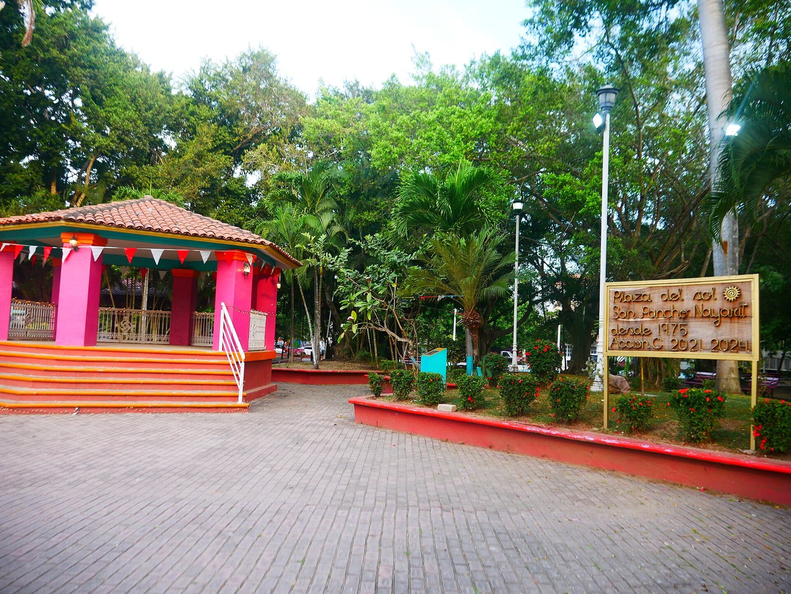 Pink gazebo with a tile roof and a wooden Plaza del Sol sign in San Pancho, surrounded by trees