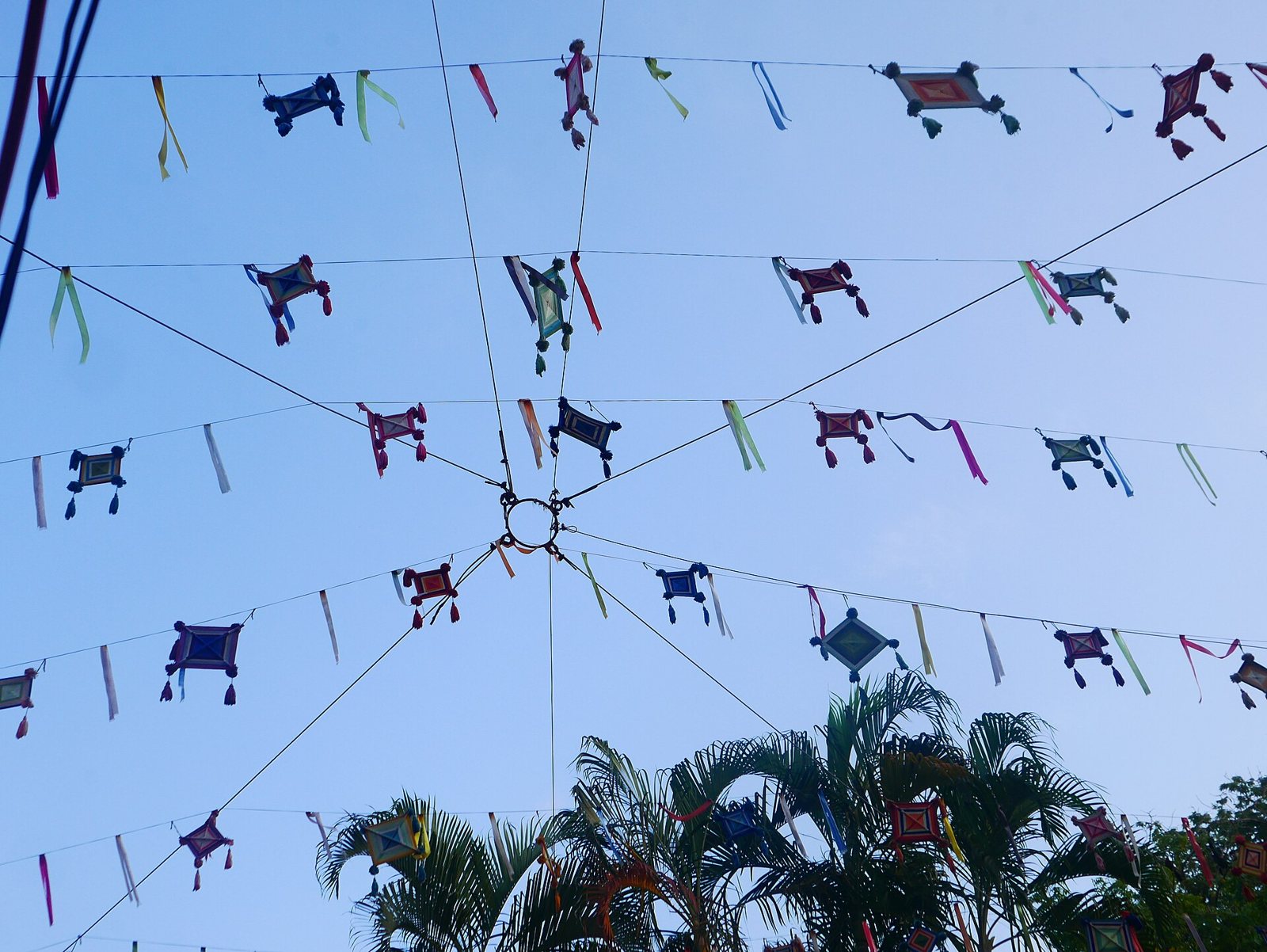 Colorful woven God's eyes (ojos de dios) strung on lines above a street in San Pancho, with palm trees and blue sky