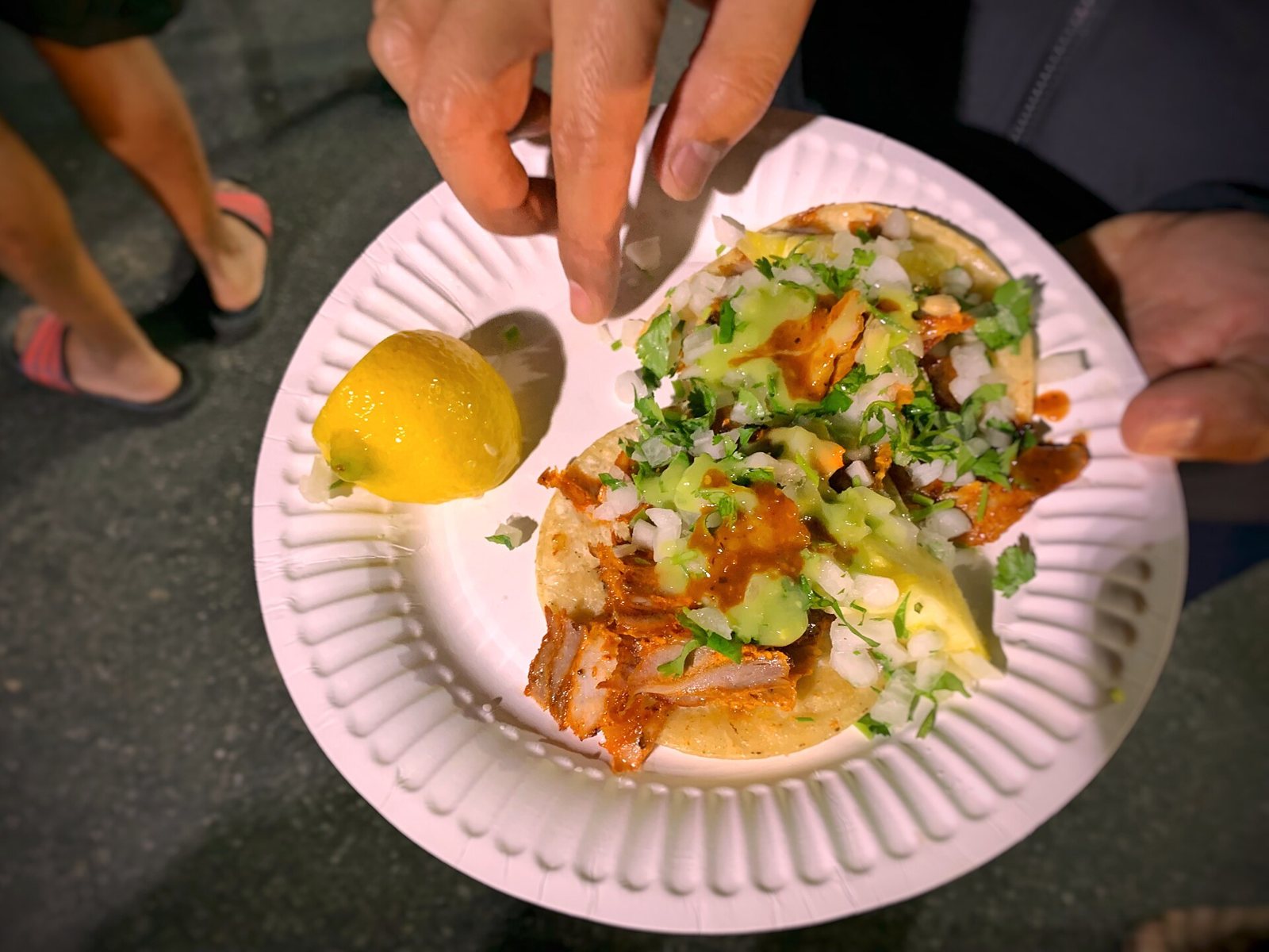 Two tacos al pastor with onion, cilantro and salsas on a paper plate, served at night on the street