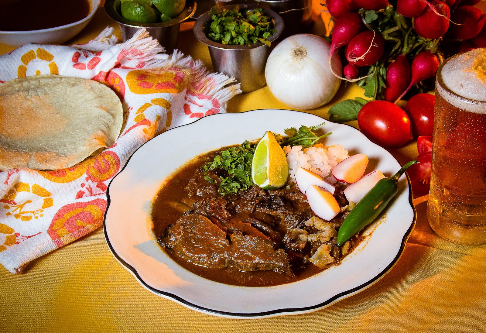 Bowl of birria topped with onion, cilantro, radish, lime and a chile, next to corn tortillas and a mug of beer