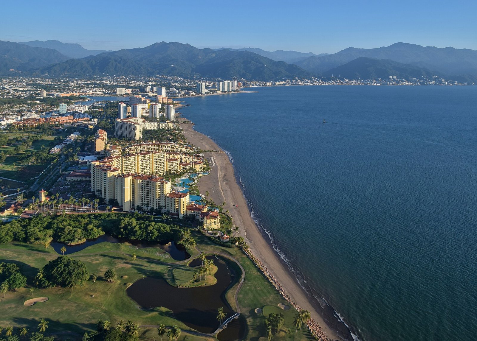 Aerial view of the Puerto Vallarta coastline with beachfront hotel towers, a golf course in the foreground and mountains behind the city
