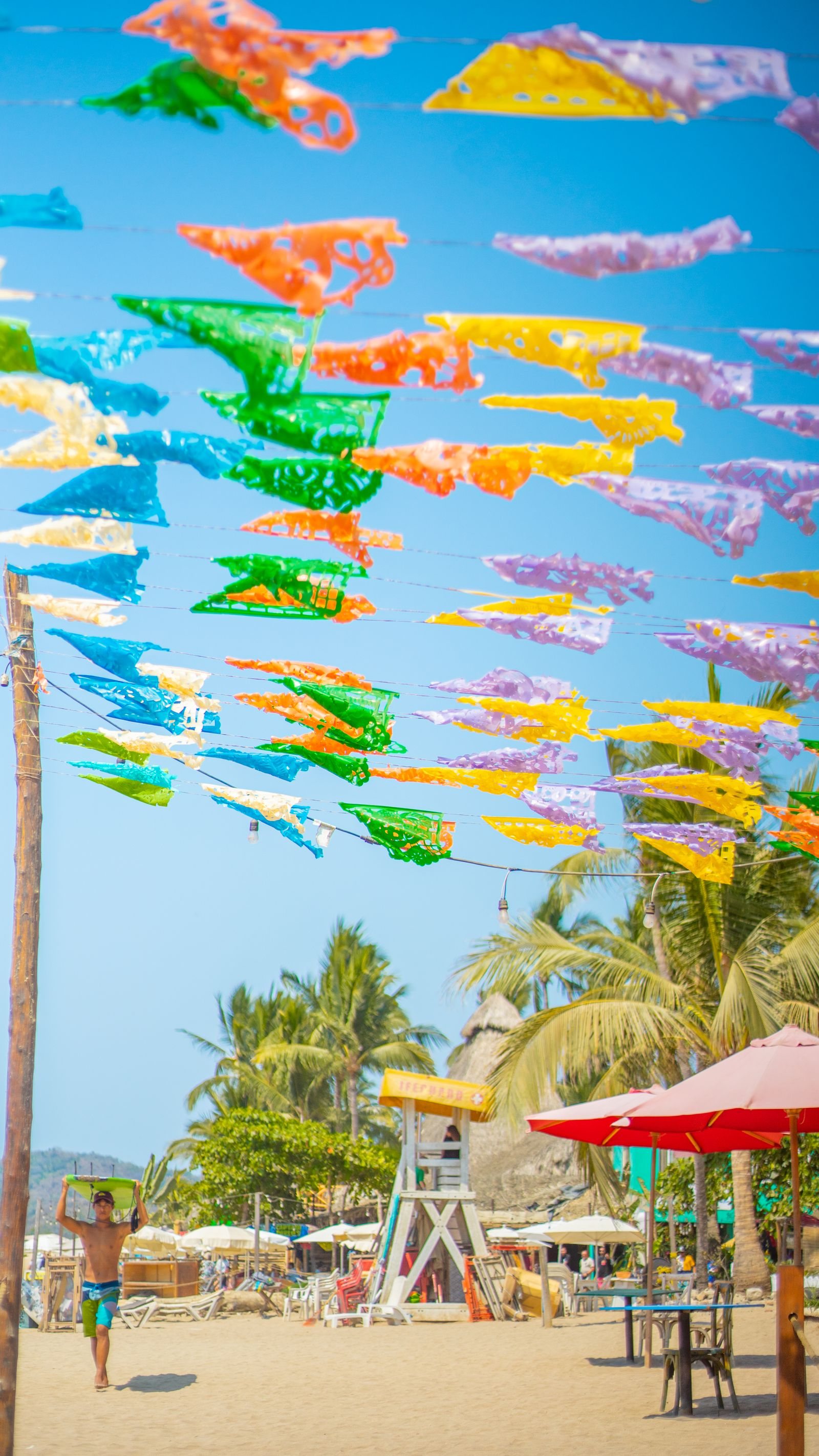 Colorful papel picado banners strung over Sayulita beach, with umbrellas, palm trees and a lifeguard tower behind