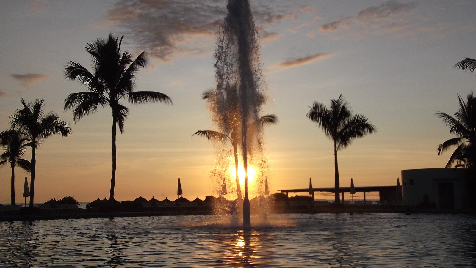 Water fountain in a pool at sunset, with silhouetted palm trees and the sun over the ocean in Nuevo Vallarta