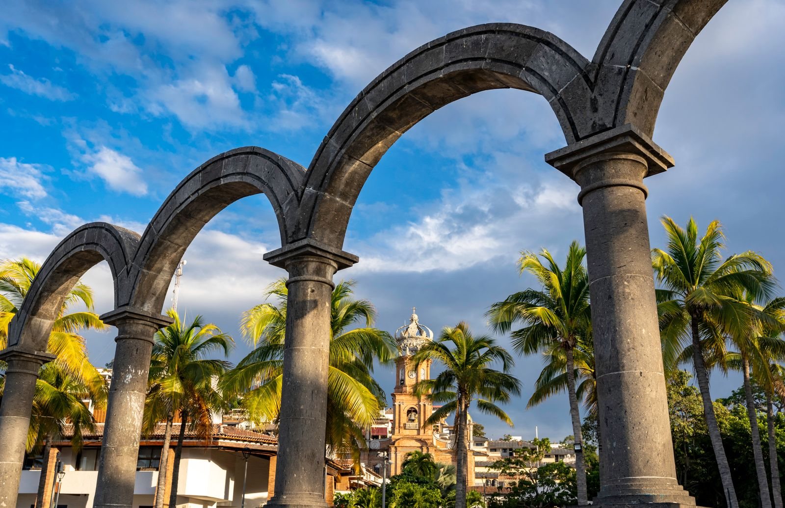 Stone arches on the Puerto Vallarta Malecon framing palm trees and the crowned tower of the Our Lady of Guadalupe church