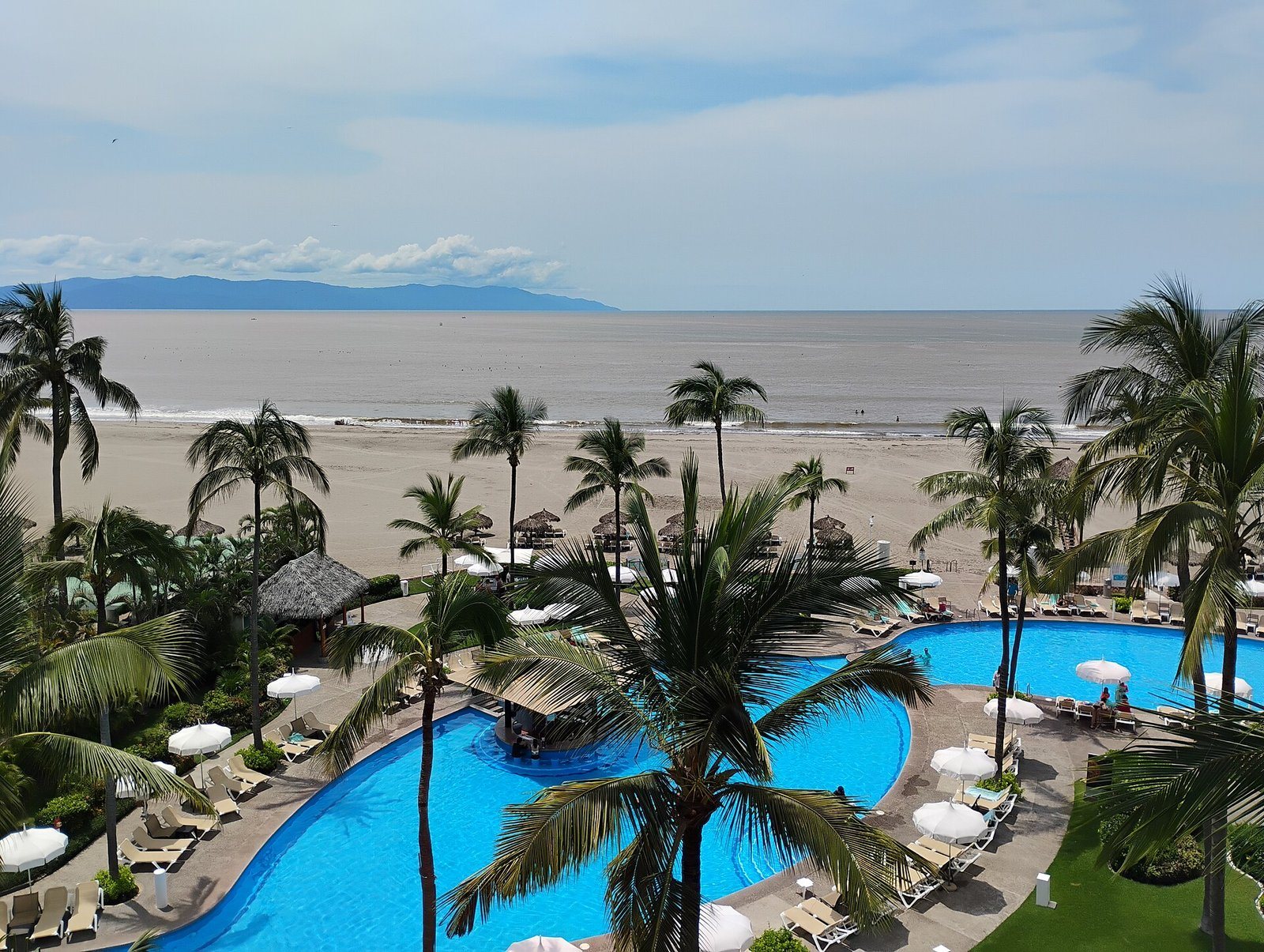 Elevated view of blue pools lined with palm trees and umbrellas, with a wide beach and Banderas Bay behind