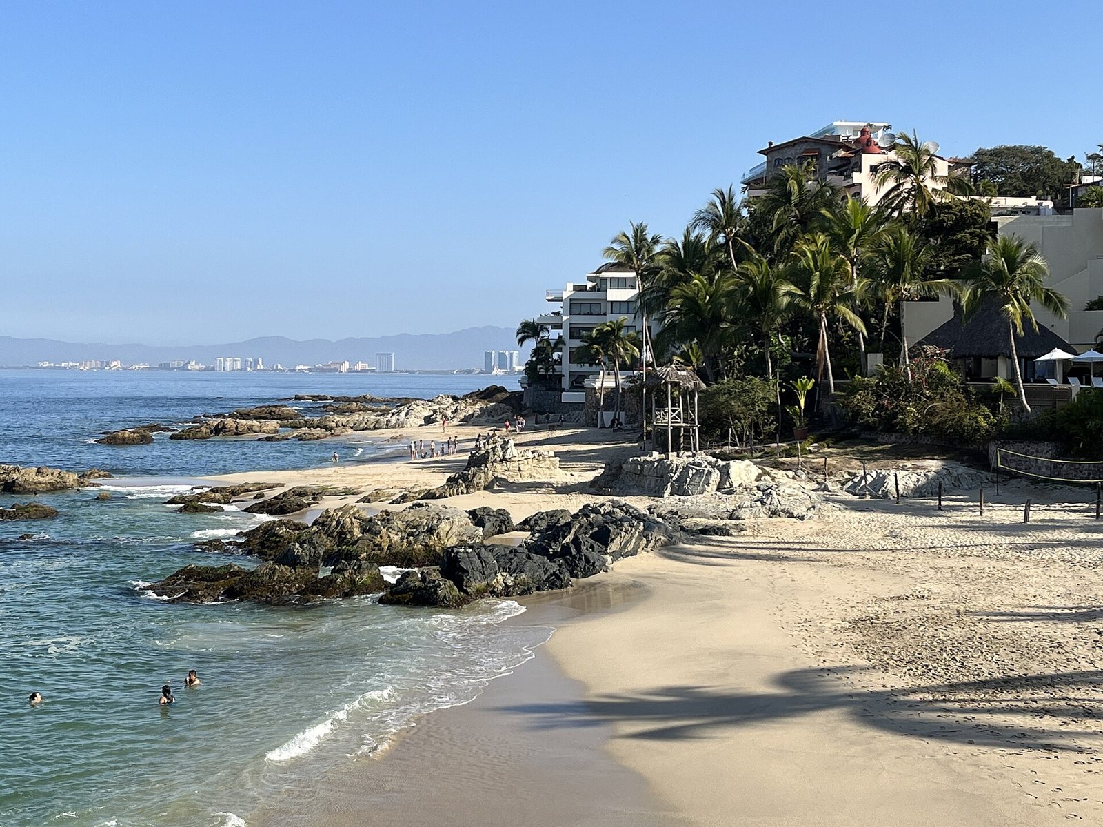 Light sandy beach with volcanic rocks, palm trees and buildings among the greenery, with the city of Puerto Vallarta in the distance