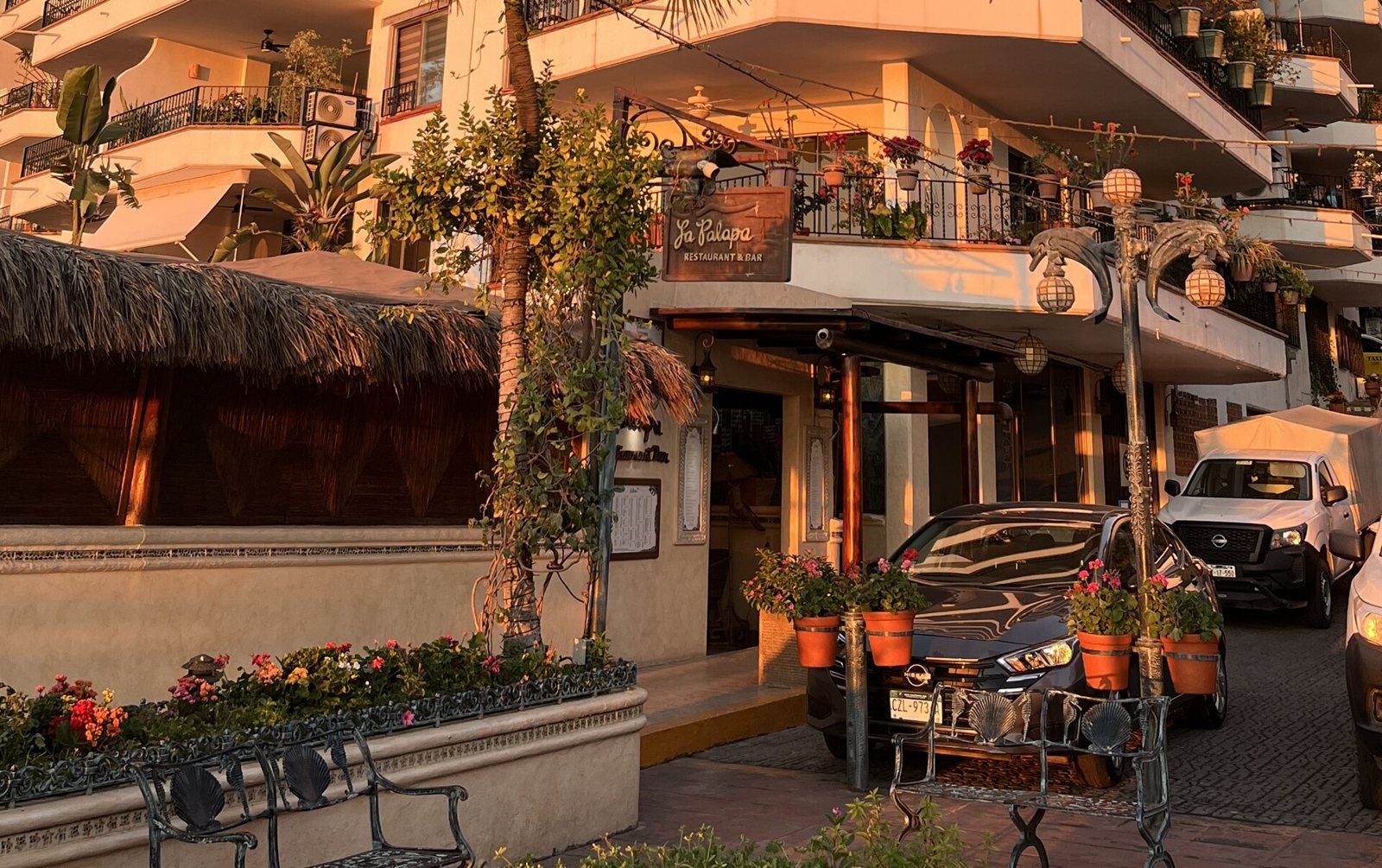 Entrance of La Palapa Restaurant & Bar with a thatched palm roof, potted flowers and golden sunset light on Púlpito Street