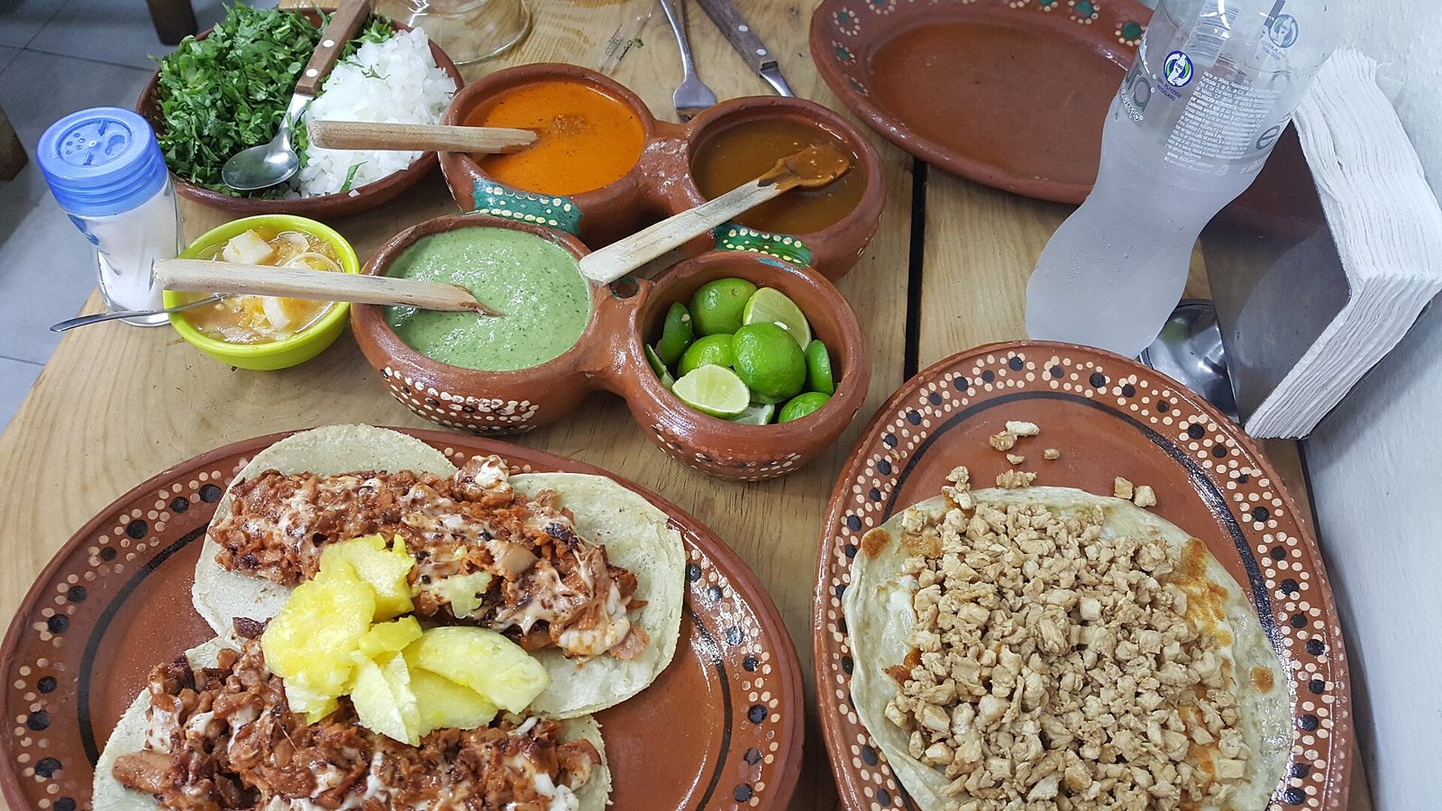 Al pastor tacos topped with pineapple on a clay plate, next to salsas, limes, cilantro and onion on a wooden table