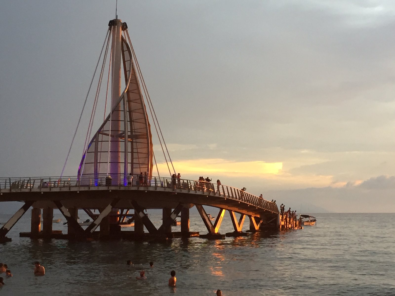 The sail-shaped Los Muertos Beach pier at sunset, with people walking on it and swimming in the sea
