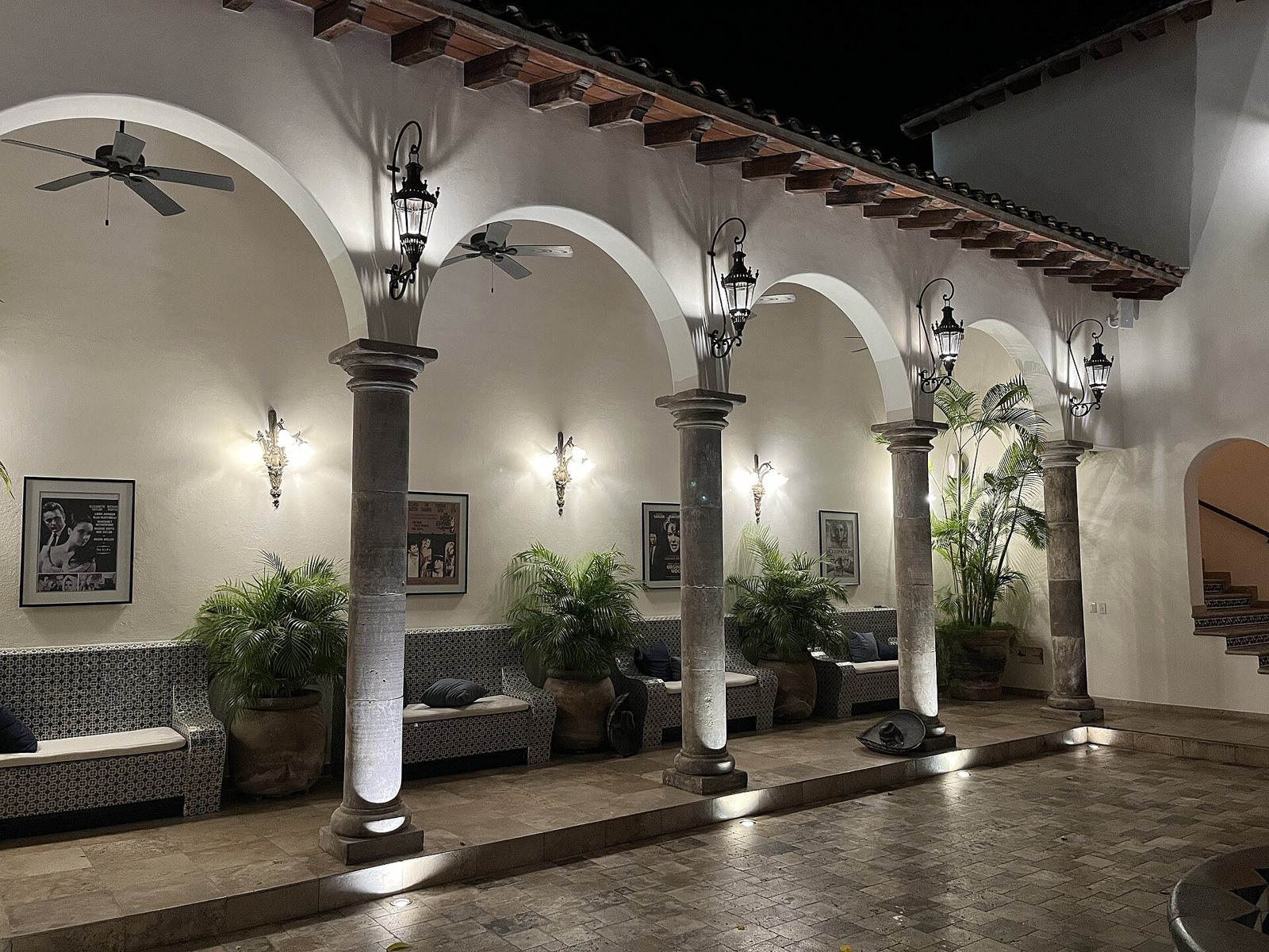 Arched portico lit at night with stone columns, lanterns, tiled benches, potted palms and framed posters of Elizabeth Taylor and Richard Burton films at Casa Kimberly