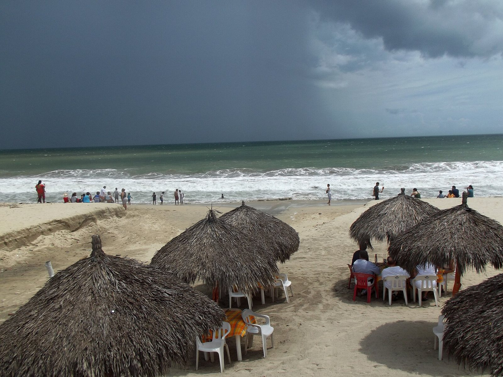 Thatched palapa umbrellas with tables and chairs on the sand in Bucerías, with swimmers at the waterline and storm clouds over the sea