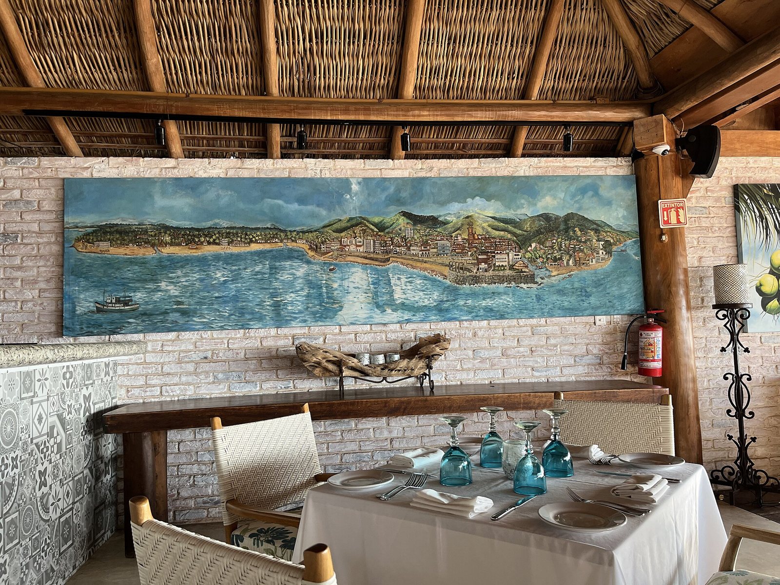Table set with a white tablecloth and blue glasses under a thatched palapa roof, with a mural of Puerto Vallarta's bay on the wall