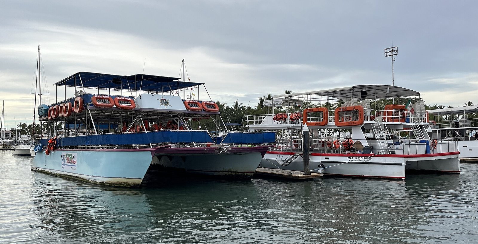 Catamarans and tour boats with orange life rings docked at a Marina Vallarta pier on a cloudy day
